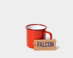 a red coffee mug sitting next to a sign that says falcon on it's side