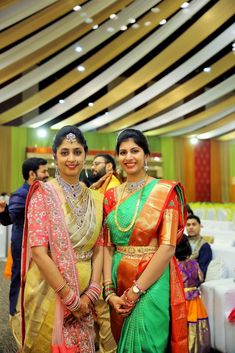 two women standing next to each other in front of a group of people wearing saris