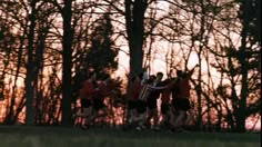 a group of young men running across a lush green field next to trees at sunset