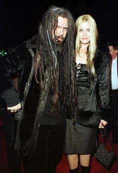 a man with dreadlocks standing next to a woman in a black dress and fur coat