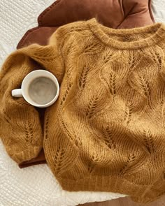 a cup of coffee sits next to a sweater on top of a bed with pillows