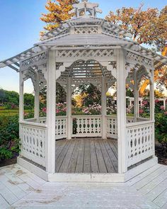 a white gazebo sitting on top of a wooden floor next to bushes and trees