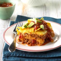 a piece of lasagna on a plate with a fork and bowl in the background