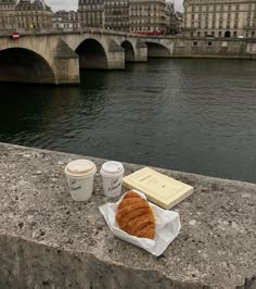 there is a croissant, coffee and book on the ledge by the water