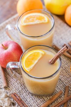 two mugs filled with orange juice and cinnamon sticks next to an apple on a table