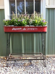 a red planter with flowers in it sitting on a metal stand next to a window