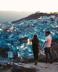 two people holding hands on top of a hill with city in the background at dusk