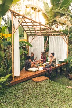 three people sitting on a wooden bench in the middle of a jungle area with white curtains