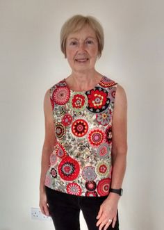 an older woman standing in front of a white wall wearing black pants and a colorful top