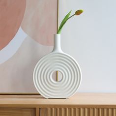 a white vase sitting on top of a wooden table next to a painting and a flower