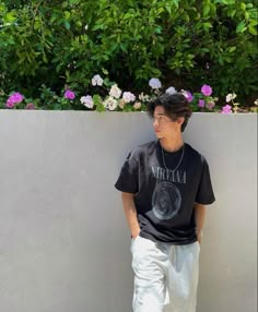 a young man leaning against a wall with his hands in his pockets, wearing white pants and a black t - shirt