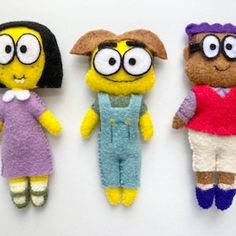 three small stuffed animals are lined up on a white surface, one is wearing overalls and the other has eyeglasses