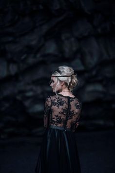 a woman standing in front of a rock wall wearing a black dress with sheer sleeves