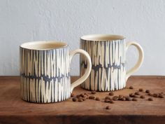 two coffee mugs sitting next to each other on top of a wooden table with coffee beans scattered around them