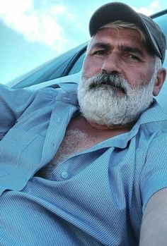 an older man with a white beard wearing a blue shirt and black hat sitting in front of a car