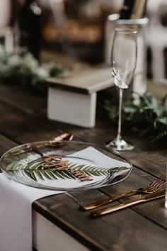 a wooden table topped with a plate covered in leaves and greenery next to wine glasses