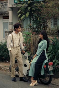 a man standing next to a woman sitting on a scooter in the street