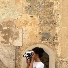 a woman holding a camera up to her face while standing in front of a stone wall