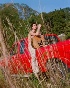 a man standing in the bed of a red pickup truck holding a guitar and wearing khaki pants