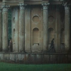 two people walking in front of an old building with many pillars and arches on the side