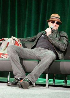 a man sitting on top of a black couch next to a green curtain holding a microphone