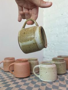 a hand is holding a coffee mug over several small cups on a checkered tablecloth