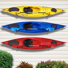 three kayaks lined up in front of a white wall with plants and shrubs around them