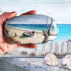 a hand holding a painted rock with a boat on the beach and seagulls flying over it