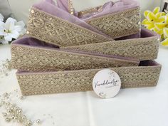 three purple and gold jewelry boxes sitting on top of a white cloth covered table with flowers in the background