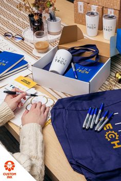 Photo of a person's hands writing gift tags on a desk that is covered in branded items like insulated mugs, notebooks, pens, and tote bags Business Merchandise, Products Ads, Company Swag, Candy Gift Box, Kit Ideas, Custom Ink, Care Packages, Holiday Gift Ideas