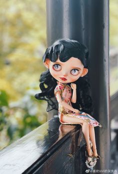 a doll sitting on top of a black pole next to a tree trunk and wearing a dress