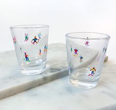 two glass cups sitting on top of a marble counter next to each other, one with colorful people painted on it