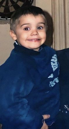 a little boy that is sitting down with a blue coat over his shoulders and smiling at the camera