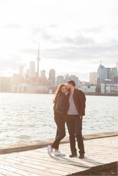 an engaged couple standing on a dock in front of the water and city skyline at sunset