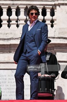 a man in a blue suit and sunglasses stands on a red carpet next to a black suitcase