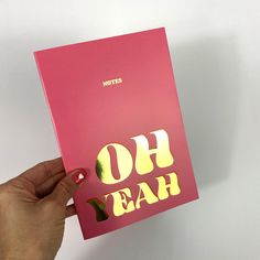 a person holding up a pink notebook with yellow lettering on it that says oh yeah