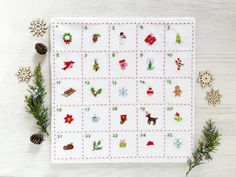 a cross stitch christmas calendar on a white table with pine cones and evergreen branches around it