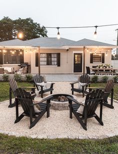 a fire pit surrounded by lawn chairs in front of a white house with string lights