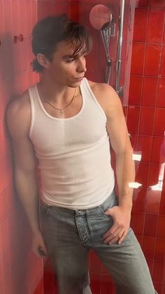 a young man leaning against a red tiled wall in front of a shower with his hands on his hips