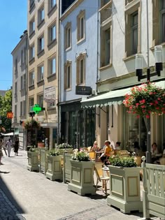 people are sitting at tables on the sidewalk in front of tall buildings with flower boxes