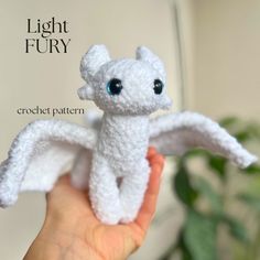 a hand holding a small white crochet dragon with big blue eyes and wings