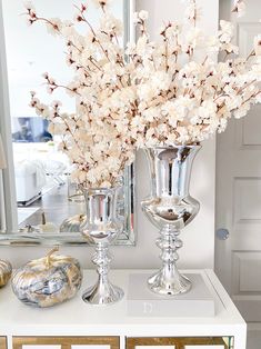 two silver vases with white flowers in them on a table next to a mirror