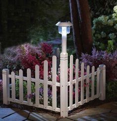 a white picket fence with a light on it in front of some flowers and trees