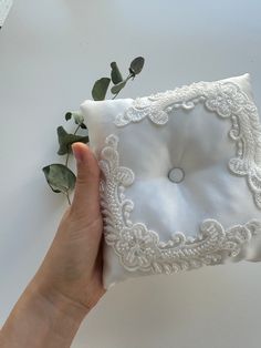 a hand holding a white pillow with lace on it and a plant in the corner