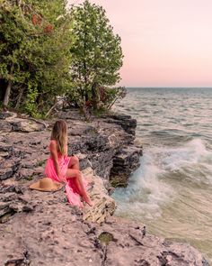 a woman in a pink dress sitting on the edge of a cliff by the ocean