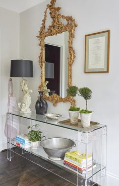 a glass console table with plants, books and a mirror on the wall above it