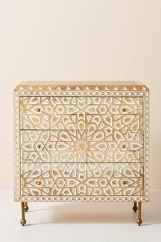 a white and gold chest of drawers with intricate carvings on the front, in an off - white room