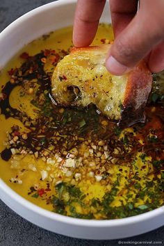 a person dipping food into a bowl of soup with bread and herbs on the side