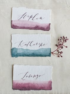 three watercolored business cards with calligraphy on them and some flowers in the background