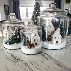 three glass jars with houses and trees in them on a marble countertop next to a christmas tree